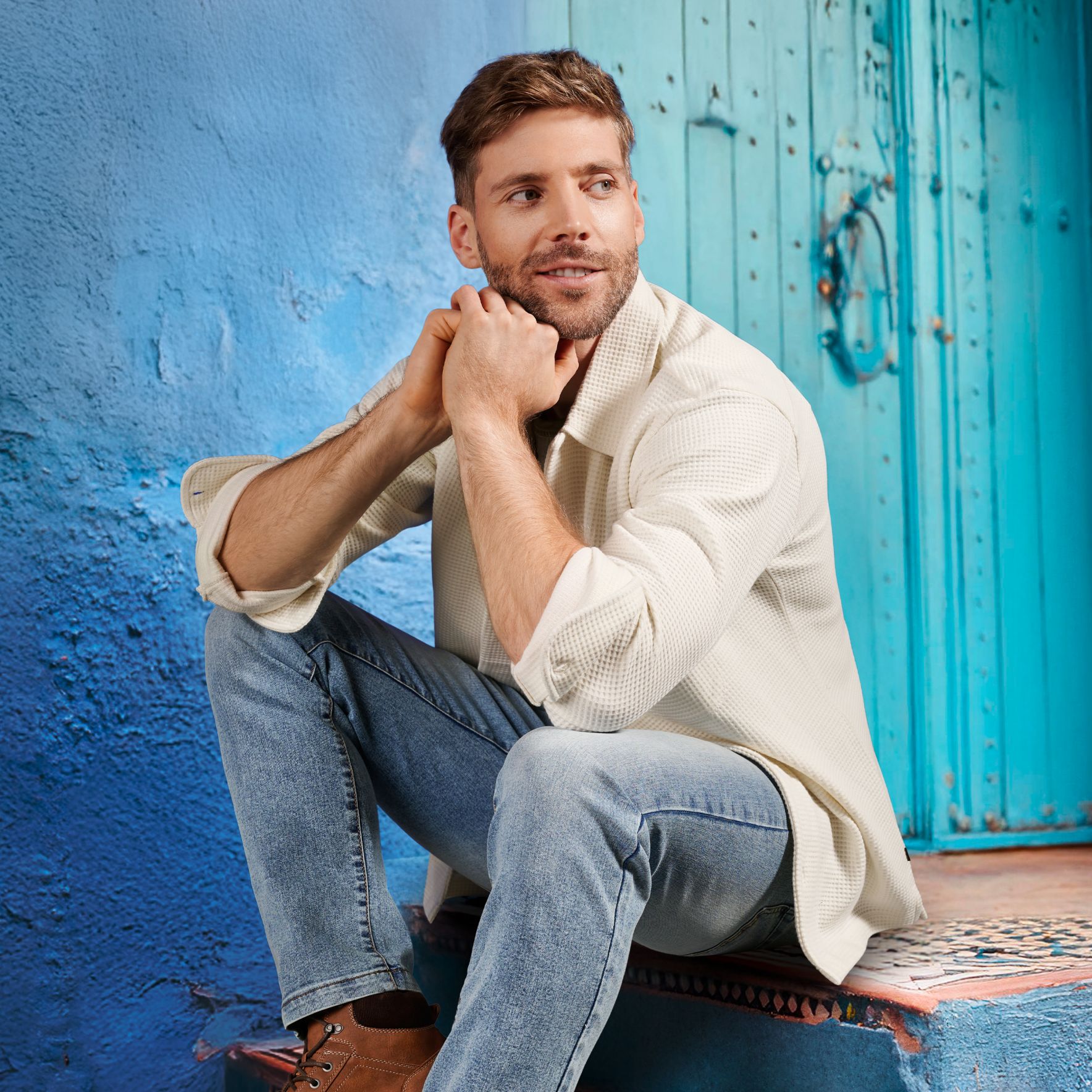 Ein Mann sitzt auf einer blauen Treppe mit Mosaikoptik. Im Hintergrund ist ein blaues Gebäude, er trägt ein weiß-beiges Hemd und eine blaue Jeans.