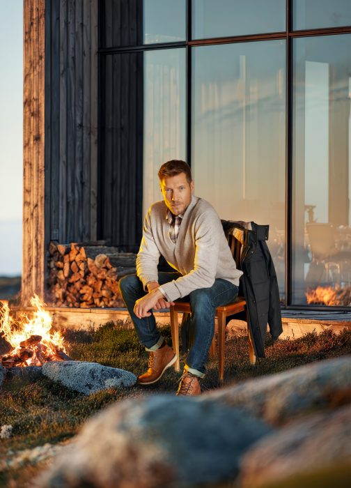 Mann in Herbstjacke sitzt entspannt vor einem Feuer bei einer Hütte