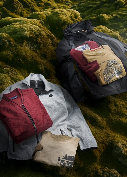 Gefaltete Herrenjacken und Pullover in Rot, Beige und Grau auf moosbewachsenem Untergrund arrangiert